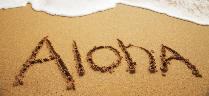 Inviting "Aloha" written in soft beach sand with surf approaching.